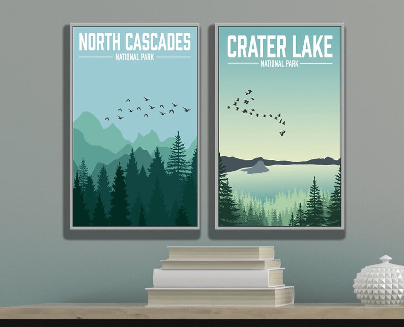 May include: Two framed posters with a minimalist design. The poster on the left features a mountain range with a flock of birds flying overhead. The text on the poster reads "North Cascades National Park". The poster on the right features a lake with a small island in the middle. The text on the poster reads "Crater Lake National Park".