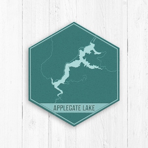 Puede incluir: Un letrero hexagonal azul verdoso con un contorno blanco y el texto "APPLEGATE LAKE" en blanco. El letrero presenta un contorno azul claro de un lago.