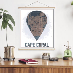 May include: A poster featuring a map of Cape Coral, Florida, in the shape of a pin. The map is in shades of blue and orange. The poster has a white background and the city name is printed in black at the bottom.