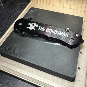 May include: A black folding knife with the text "THE WOODSMAN" and a skull graphic. The knife is resting on a black surface inside a gold-colored machine with a fan.