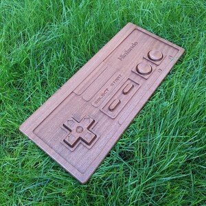 May include: A wooden replica of a classic Nintendo controller. The controller is brown and has the Nintendo logo and the words "Select" and "Start" printed on it. The controller is lying on a bed of green grass.