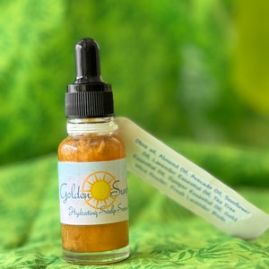 May include: A small glass bottle of Golden Sun Hydrating Scalp Serum with a black dropper. The label features a sun graphic and the product name. A white ingredient list is next to the bottle. The serum is a golden color.