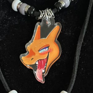 Puede incluir: Dos collares con colgantes de personajes de dibujos animados. El collar superior presenta un colgante de una criatura naranja y roja con forma de dragón con la lengua fuera. El collar inferior tiene un colgante de una criatura con forma de gato con un cuerno y la lengua fuera.