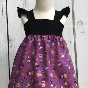 Può includere: Abito viola senza maniche con spalline nere e maniche a sbuffo. L'abito ha una stampa di personaggi dei Peanuts vestiti con costumi di Halloween. La stampa include il testo "Boo!" e "Trick or Treat".