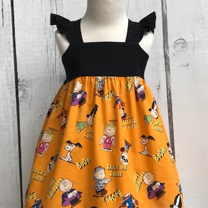 Può includere: Un vestito giallo e nero senza maniche con una stampa di Halloween dei Peanuts. La stampa presenta personaggi come Snoopy, Charlie Brown e Lucy vestiti con costumi di Halloween. Il vestito ha un corpetto nero e una gonna gialla con un bordo a balze.