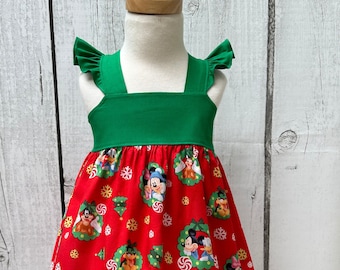 May include: A red and green Christmas dress with a Mickey Mouse and Minnie Mouse print. The dress has a green bodice and a red skirt with a ruffle trim on the straps.