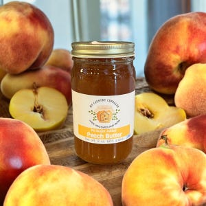 May include: A glass jar of peach butter with a gold lid. The jar is labeled "My Country Cupboard" and "No Sugar Added Peach Butter". The jar is surrounded by fresh peaches.
