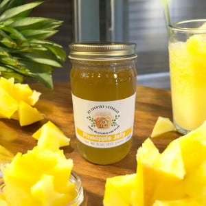 May include: A glass jar of pineapple jelly with a gold lid. The jar has a white label with the text "My Country Cupboard" and "Pineapple Jelly". The label also includes the text "Jams, Mustards and More" and "Net Wt. 8 oz. (255 grams)".