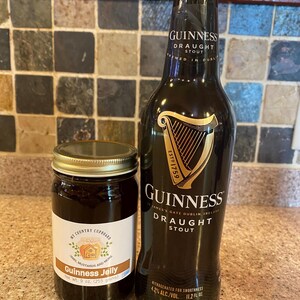 May include: A bottle of Guinness Draught Stout next to a jar of Guinness Jelly. The stout bottle is dark with gold lettering and a harp logo. The jelly jar has a white label with text and a gold lid. The jelly jar is labeled as 9 oz.