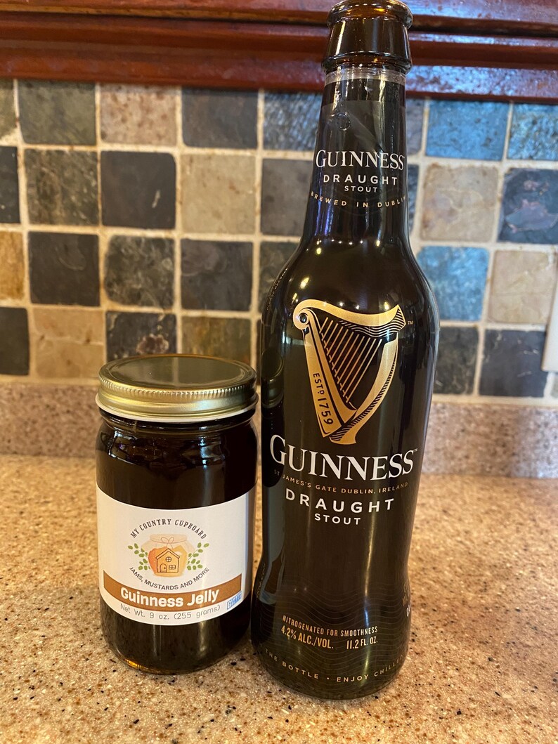 May include: A bottle of Guinness Draught Stout next to a jar of Guinness Jelly. The stout bottle is dark with gold lettering and a harp logo. The jelly jar has a white label with text and a gold lid. The jelly jar is labeled as 9 oz.