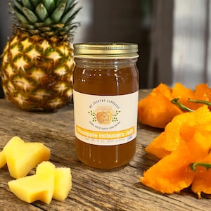 May include: A glass jar of pineapple habanero jelly with a gold lid. The jar is labeled "My Country Cupboard" and "Pineapple Habanero Jelly". The jar is sitting on a wooden surface with pineapple and habanero pepper slices.