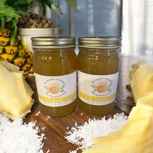 May include: Two glass jars filled with a yellow, translucent jam labeled "Pina Colada Jam" with a net weight of 9 oz (255 grams). The jars are on a wooden surface with pieces of pineapple and shredded coconut.