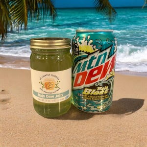 May include: A glass jar of green Baja Dew Jelly with a label that reads "My Country Cupboard Jams, Mustards and More Baja Dew Jelly Net Wt. 9 oz. (255 grams)" next to a can of Mountain Dew Baja Blast.