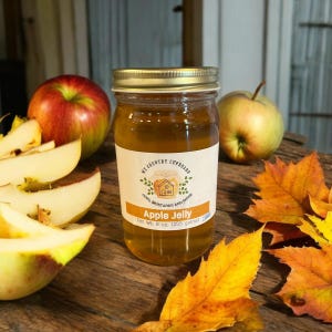 May include: A glass jar of apple jelly with a gold lid. The jar has a label that says "My Country Cupboard" and "Apple Jelly". The label also has a small illustration of a house with a chimney and a red roof. The jar is sitting on a wooden surface with fall leaves and apples.