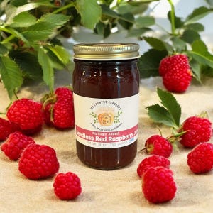 May include: A glass jar of seedless red raspberry jam with a white label that reads "My Country Cupboard" and "Seedless Red Raspberry Jam, No Sugar Added, Net Wt. 9 oz. (255 grams)". The jar is surrounded by fresh red raspberries.
