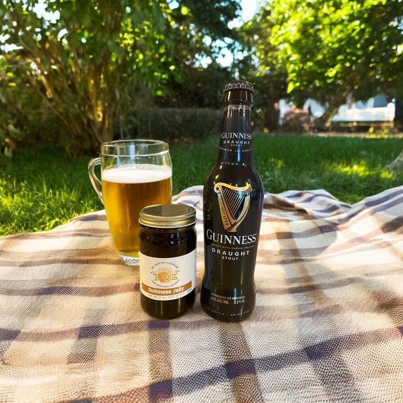 May include: A glass mug of beer, a jar of Guinness Jelly, and a bottle of Guinness Draught Stout sit on a plaid blanket outdoors. The Guinness bottle is dark with a gold harp logo. The jelly jar has a label.