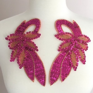 May include: Two magenta sequined appliques, each featuring a stylized floral design. The appliques have a gold beaded outline and accents, with a light pink mesh backing. These decorative embellishments are suitable for clothing or craft projects.