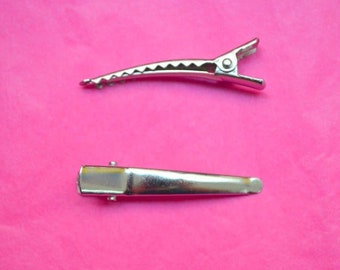 May include: Two silver-colored metal alligator hair clips on a bright pink background. The clips are open, showing their teeth-like grip. One clip is positioned above the other.