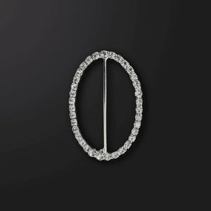 Puede incluir: Una hebilla ovalada plateada con un diseño de diamantes de imitación transparentes. La hebilla tiene una barra de metal que atraviesa el centro.