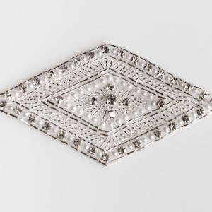 May include: A silver-toned, diamond-shaped embellishment with a detailed pattern. The design features rows of small, white pearls and clear, sparkling rhinestones. The edges are outlined with a silver border, creating a decorative accessory.