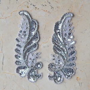 May include: Two silver sequined fabric appliques in the shape of a stylized leaf. The appliques are on a white background.