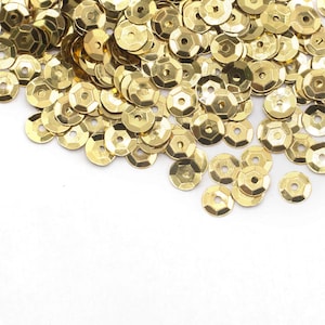 May include: A pile of gold-coloured sequins, scattered on a white surface. The sequins are round with a flat back and a raised, faceted front.