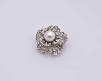 May include: A silver-toned flower-shaped brooch with a central pearl and encrusted with small, clear crystals. The brooch has a detailed, layered petal design and a secure pin backing.