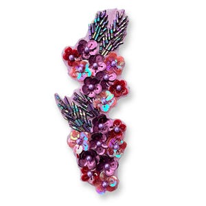 May include: A decorative floral applique in shades of pink, red, and purple. The applique features sequined flowers and beaded accents, creating a textured and dimensional design. The base is a light pink fabric.