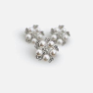 Puede incluir: Un conjunto de tres pendientes de perlas y pedrería. Los pendientes están dispuestos en un diseño floral, con un grupo central de pedrería rodeado de seis perlas de imitación. Los pendientes tienen un engaste de metal plateado.