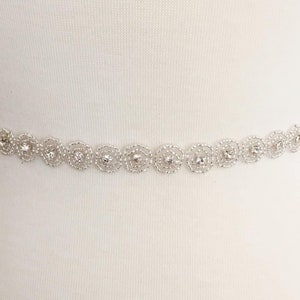May include: A delicate silver-colored belt featuring a repeating floral design. Each flower is crafted with a circular, openwork pattern and a central, sparkling rhinestone. The belt is shown against a white background.