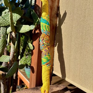 May include: A handcrafted, yellow wooden dowel wrapped with colorful yarn. The yarn includes sections of turquoise, green, and a mix of orange, purple, and yellow. The dowel is partially visible at the top and bottom.