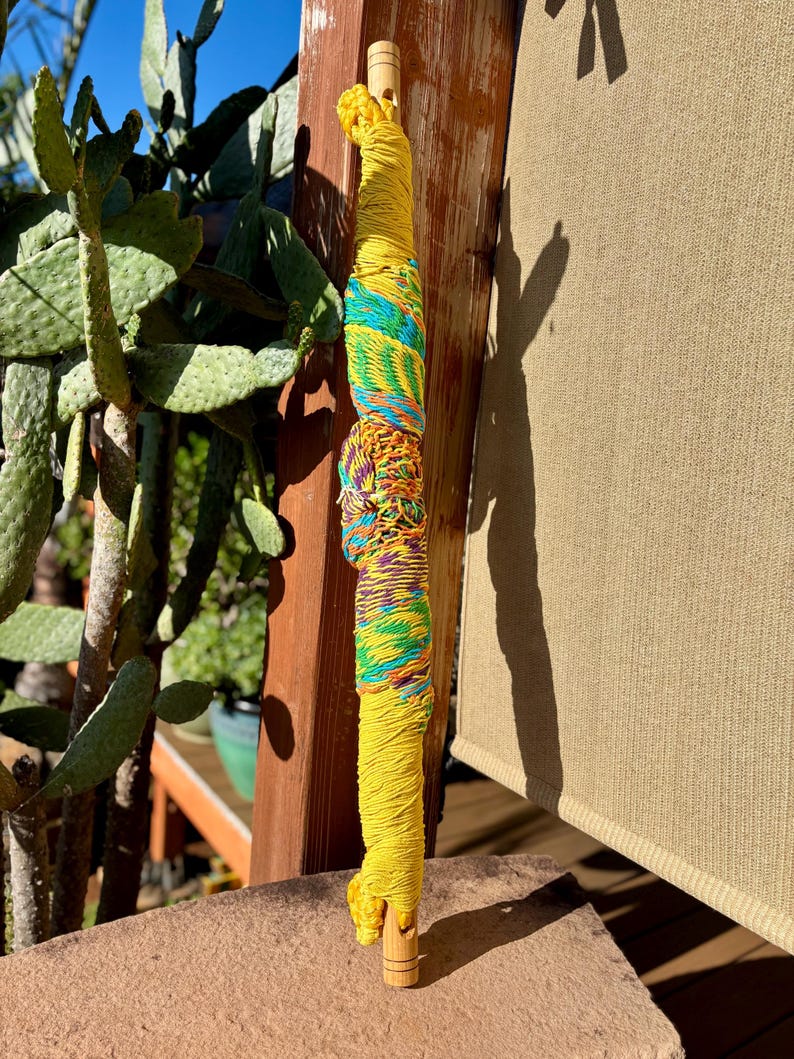 May include: A handcrafted, yellow wooden dowel wrapped with colorful yarn. The yarn includes sections of turquoise, green, and a mix of orange, purple, and yellow. The dowel is partially visible at the top and bottom.