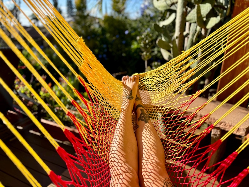 May include: A close-up shot of a yellow and red hammock. The hammock is made of woven rope and is suspended outdoors. The image shows a person's legs resting in the hammock, with tattoos visible on the legs. The background is blurred, with greenery and sunlight.