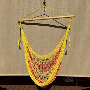May include: A yellow hammock chair with red stripes, suspended from a wooden bar and rope. The chair is made of woven material and hangs against a neutral-toned background. The hammock chair is designed for relaxation and outdoor use.