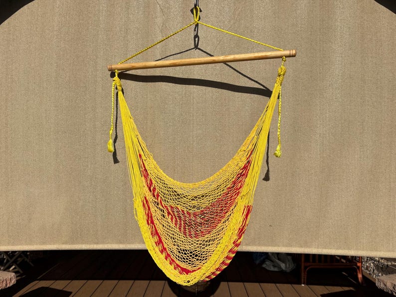 May include: A yellow hammock chair with red stripes, suspended from a wooden bar and rope. The chair is made of woven material and hangs against a neutral-toned background. The hammock chair is designed for relaxation and outdoor use.