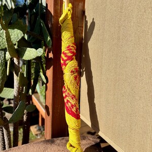 May include: A decorative bottle stopper wrapped in yellow and red yarn. The yarn is wrapped around a wooden dowel, with a wooden base. The stopper is standing upright on a stone surface, with a cactus and wooden post in the background.