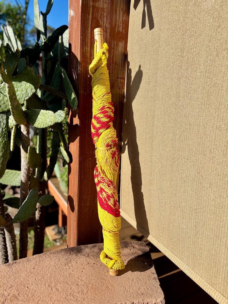 May include: A decorative bottle stopper wrapped in yellow and red yarn. The yarn is wrapped around a wooden dowel, with a wooden base. The stopper is standing upright on a stone surface, with a cactus and wooden post in the background.
