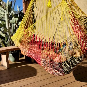 May include: A yellow, red, and black hammock hanging outdoors. The hammock is made of woven material and is suspended from a wooden structure. The hammock is empty, and the background includes a wooden deck, a small table, and plants.