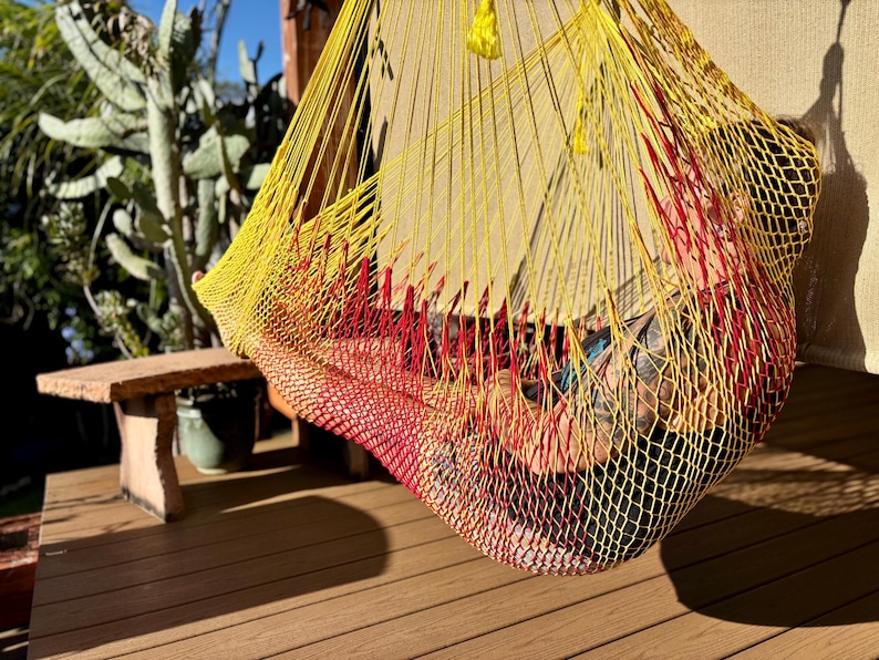 May include: A yellow, red, and black hammock hanging outdoors. The hammock is made of woven material and is suspended from a wooden structure. The hammock is empty, and the background includes a wooden deck, a small table, and plants.