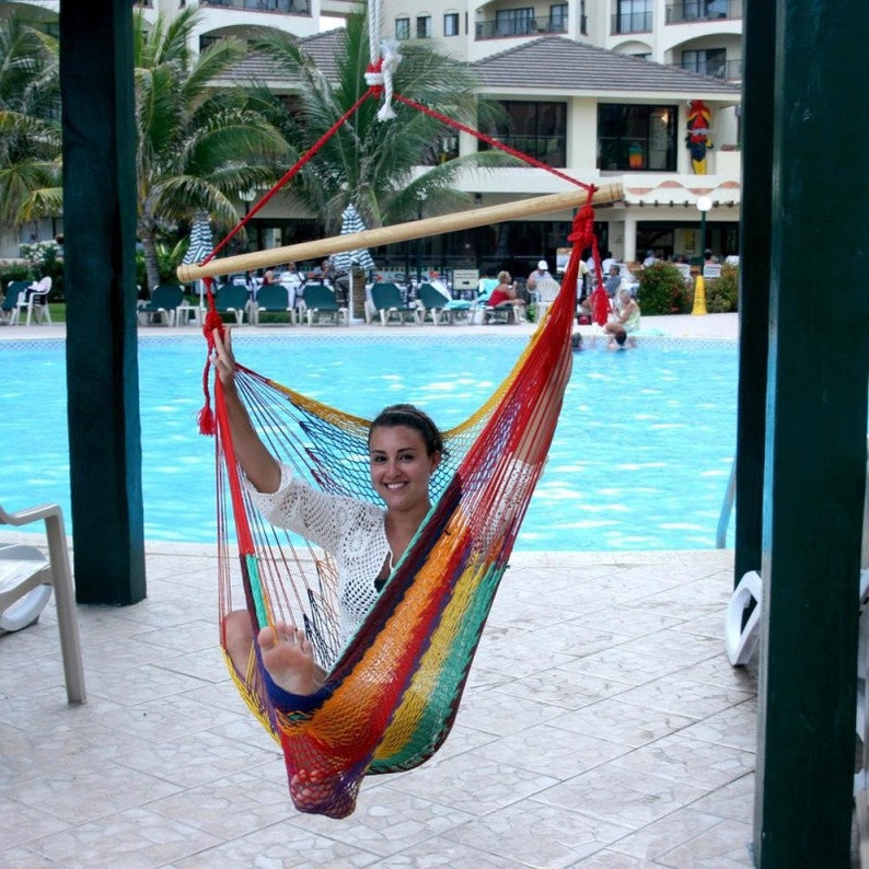 May include: A colorful hammock chair with a red, yellow, and blue striped pattern. The hammock is suspended from a wooden bar and red ropes. The background includes a swimming pool and a resort.