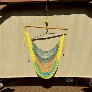 May include: A colorful hammock chair with a wooden spreader bar, suspended from a wooden structure. The hammock features a vibrant gradient of yellow, green, orange, and blue. The background is a neutral-toned fabric.