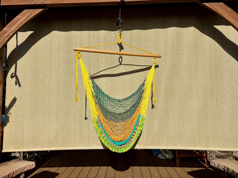 May include: A colorful hammock chair with a wooden spreader bar, suspended from a wooden structure. The hammock features a vibrant gradient of yellow, green, orange, and blue. The background is a neutral-toned fabric.