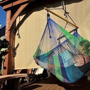 May include: A colorful hammock chair with blue, green, and orange woven netting hangs from a wooden beam. The hammock is suspended from a wooden beam and rope. A person is sitting inside the hammock. The background includes a cactus and a wooden structure.