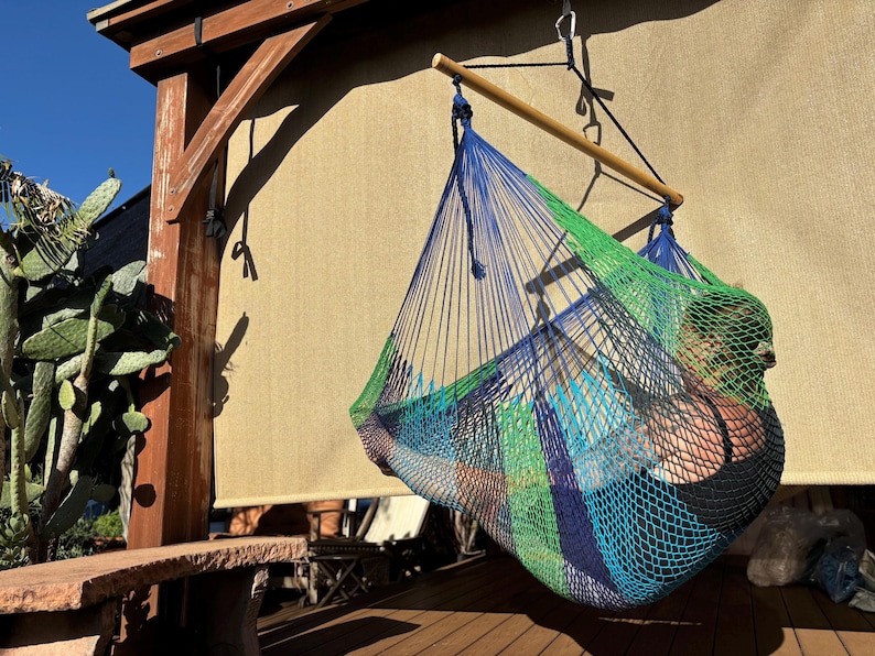 May include: A colorful hammock chair with blue, green, and orange woven netting hangs from a wooden beam. The hammock is suspended from a wooden beam and rope. A person is sitting inside the hammock. The background includes a cactus and a wooden structure.