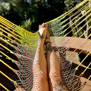 May include: A yellow and green hammock with a woven net design. The hammock is suspended, with the legs of a person resting on it. The legs have tattoos. The background is blurred with sunlight.