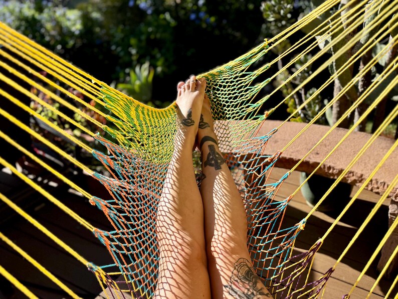 May include: A yellow and green hammock with a woven net design. The hammock is suspended, with the legs of a person resting on it. The legs have tattoos. The background is blurred with sunlight.