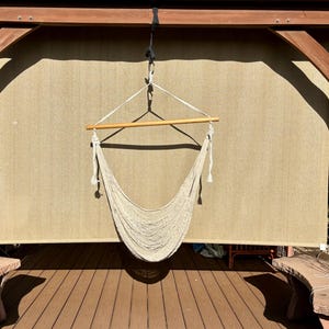 May include: A beige macrame hammock chair suspended from a wooden frame. The chair hangs in front of a beige backdrop, with a wooden beam at the top. The scene is outdoors, with a wooden deck and stone benches visible.