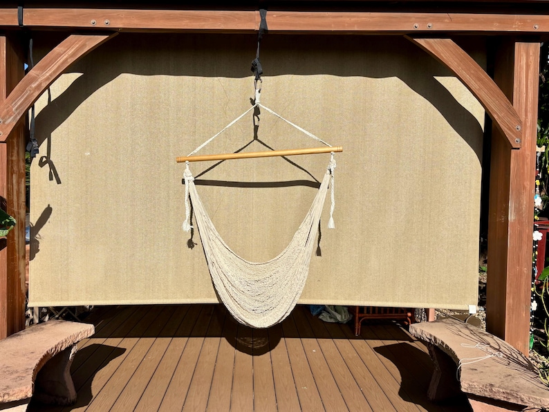 May include: A beige macrame hammock chair suspended from a wooden frame. The chair hangs in front of a beige backdrop, with a wooden beam at the top. The scene is outdoors, with a wooden deck and stone benches visible.