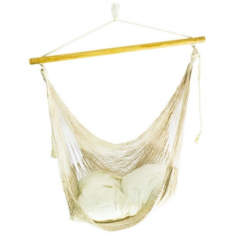 May include: A hanging hammock chair made of woven beige material, suspended from a wooden bar and white rope. Two cream-colored pillows rest inside the hammock. The chair is designed for relaxation and leisure.