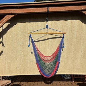 May include: A colorful hammock chair with a red, green, and blue net design, suspended from a wooden beam. The chair hangs in front of a beige backdrop, with a wooden deck below.